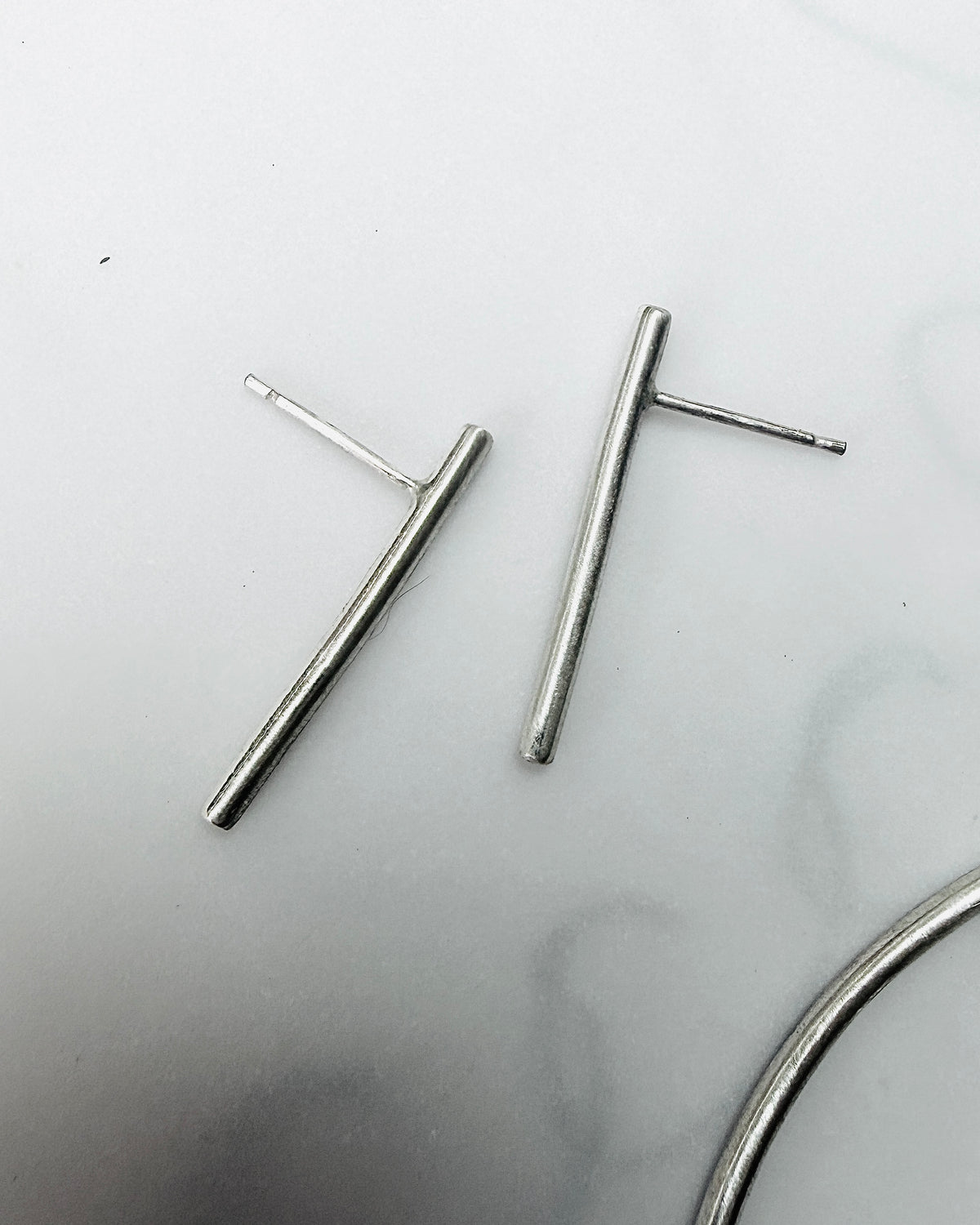 A pair of minimalist silver bar stud earrings with post backs, displayed on a light gray marble surface.
