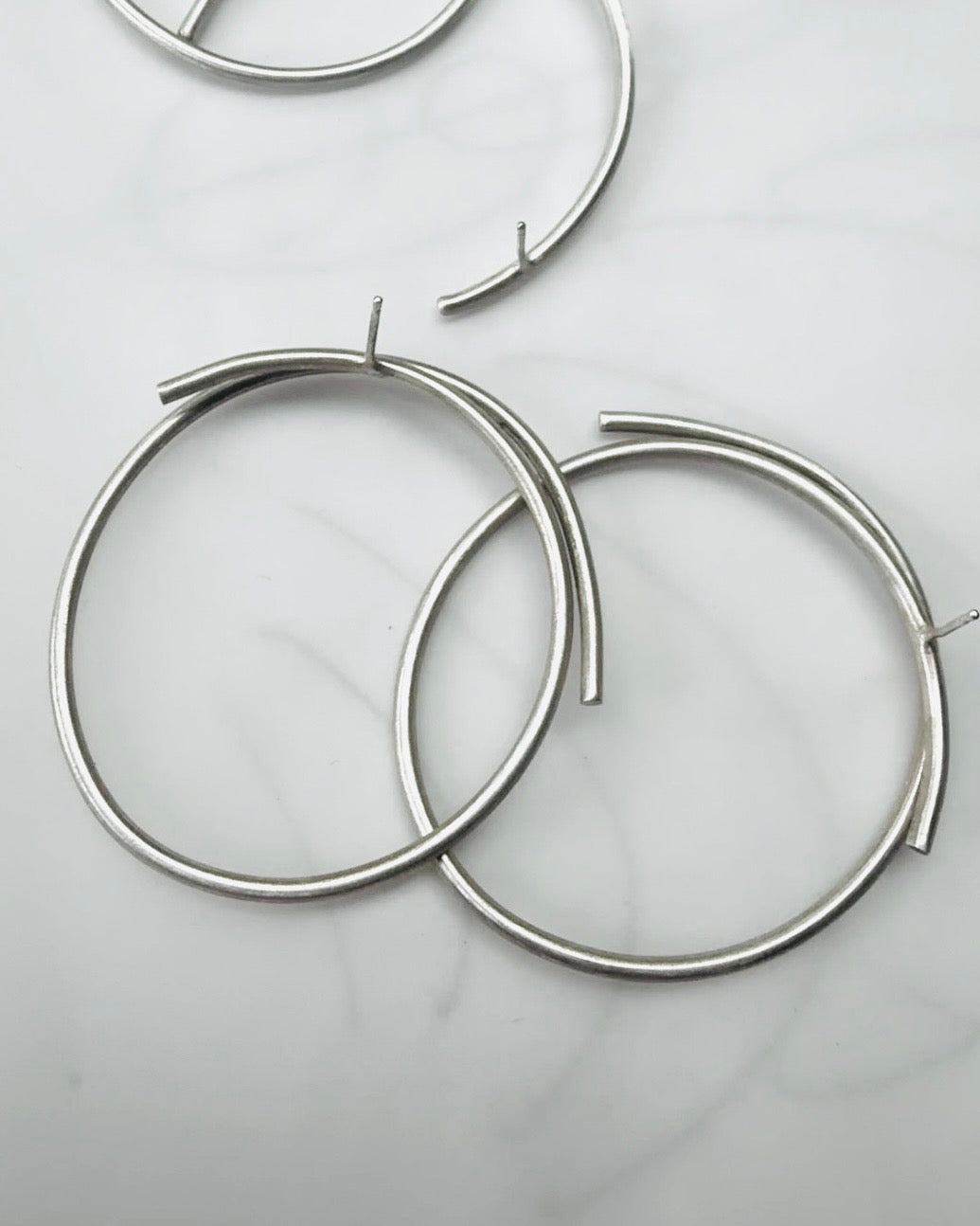 Two large silver-tone open hoop earrings with a curved bar detail at the top, displayed on a light marble surface.