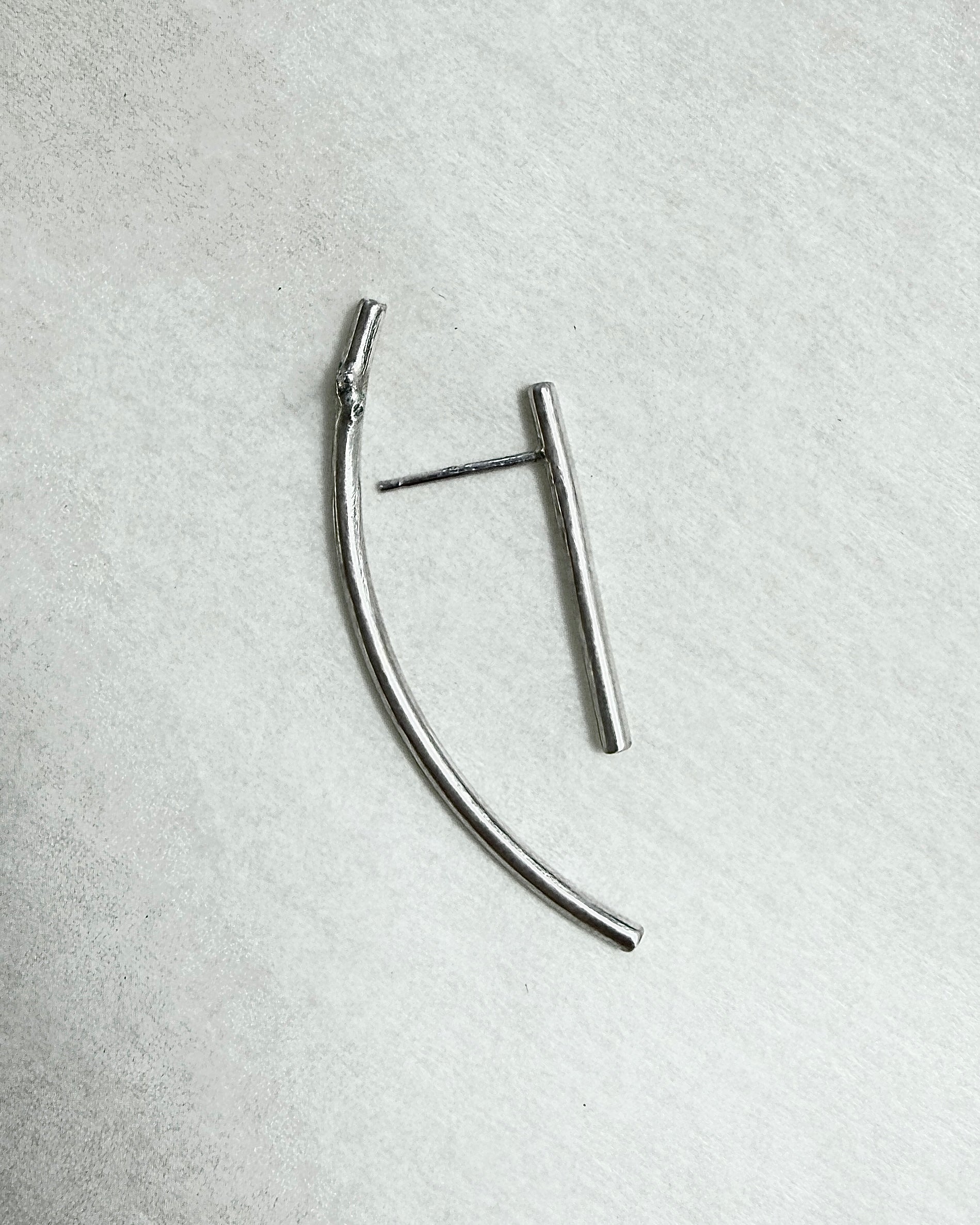 A pair of minimalist silver-toned metal earrings on a textured light gray background. One earring is a curved bar, and the other is a straight bar with a post.