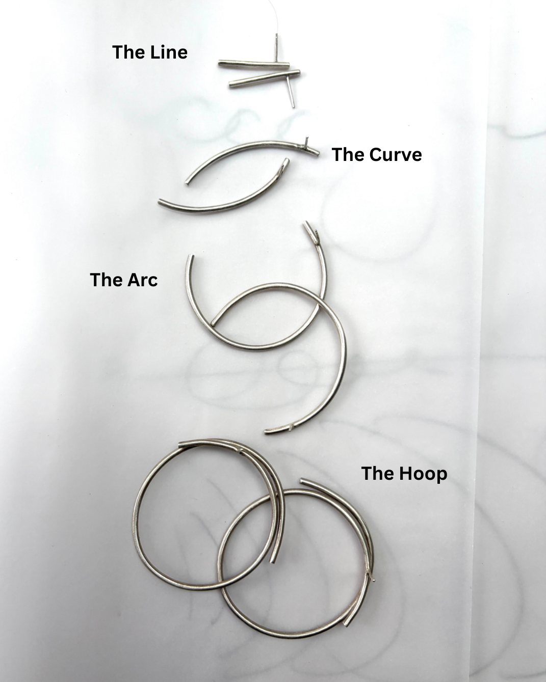 Four pairs of thin silver-toned metal hoop-style earrings arranged vertically on a white background, with handwritten labels reading 'The Line,' 'The Curve,' 'The Arc,' and 'The Hoop.'.