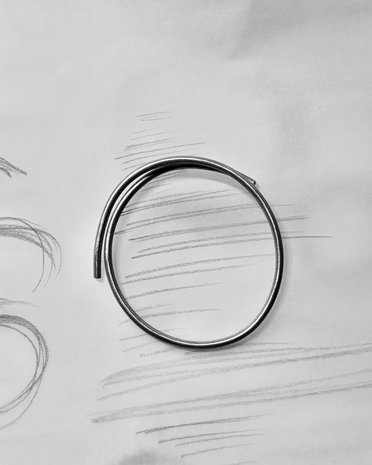 A thin, polished silver metal ring with a unique curved, looped design, resting on a light gray textured paper background with faint pencil sketches.