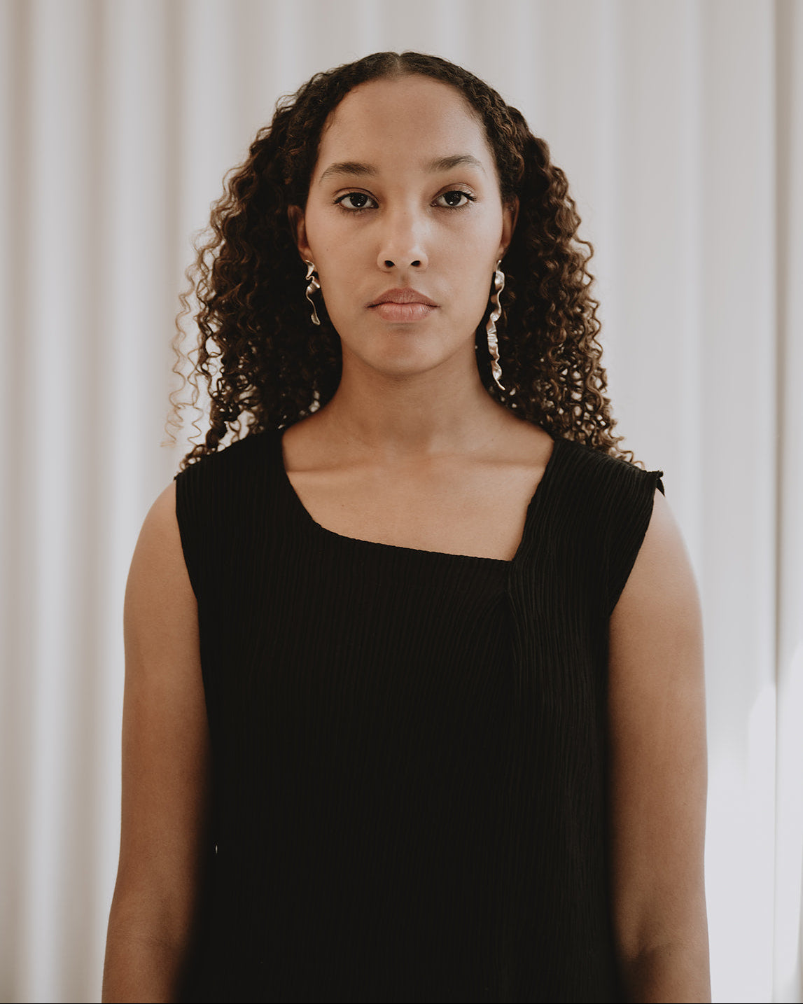 A woman with curly brown hair wearing a black sleeveless top, standing against a soft white curtain background.