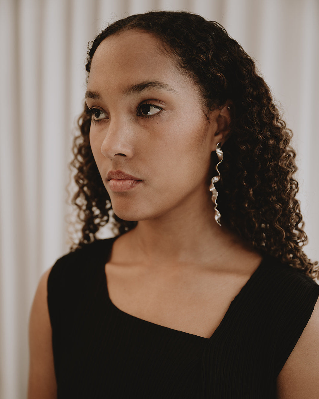 A woman with curly dark hair wearing a black sleeveless top and dangling silver earrings, photographed against a soft beige curtain background.
