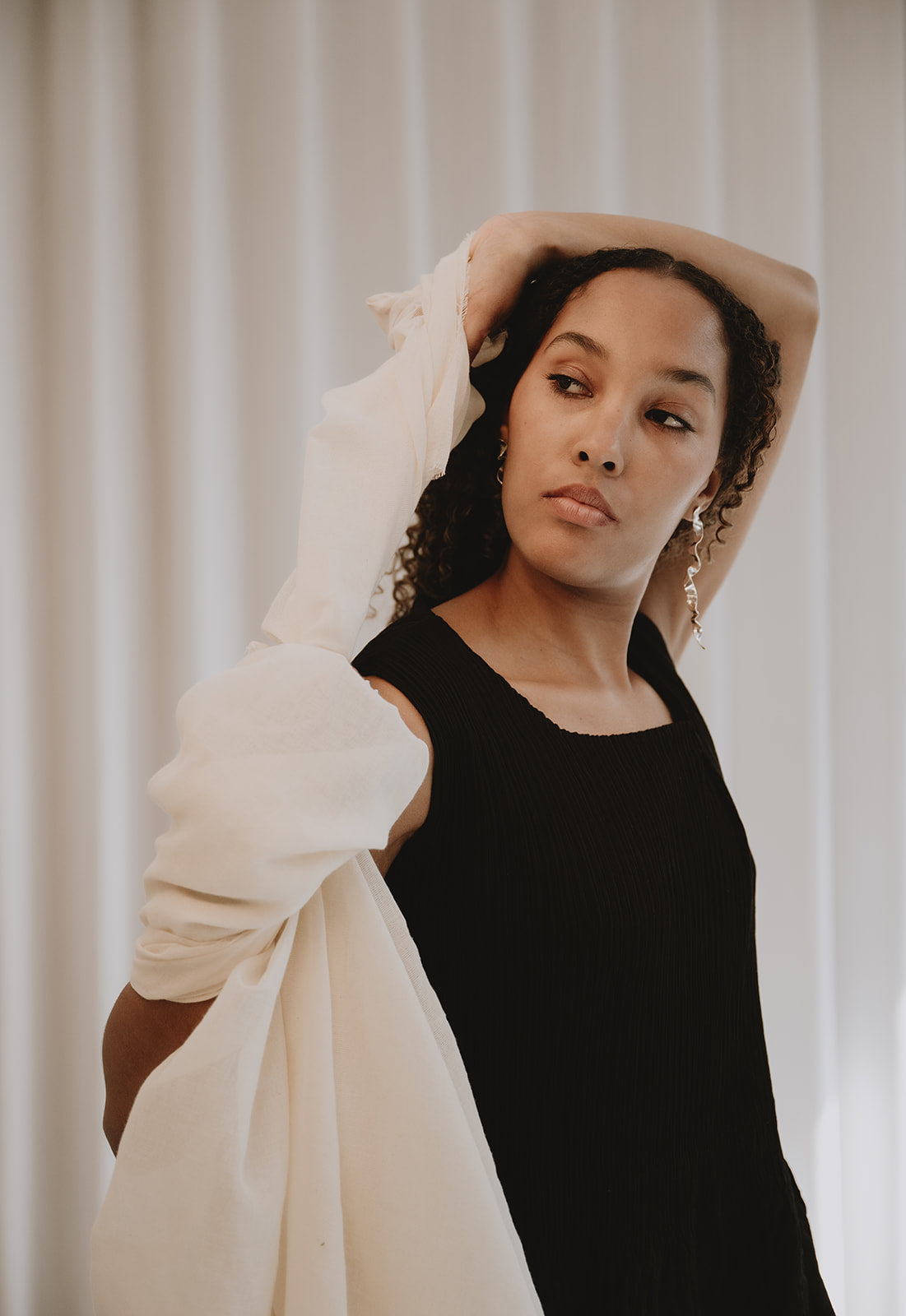 A woman posing in a black sleeveless top and holding a voluminous, off-white draped fabric or shawl against a soft, light beige curtain background.